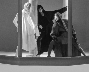Three models in hooded outfits posing inside an architectural set with columns for AnOther Magazine.