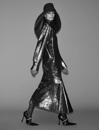 Model in dark wide-brimmed hat and metallic long dress walking against a grey backdrop