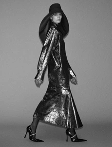 Model in dark wide-brimmed hat and metallic long dress walking against a grey backdrop
