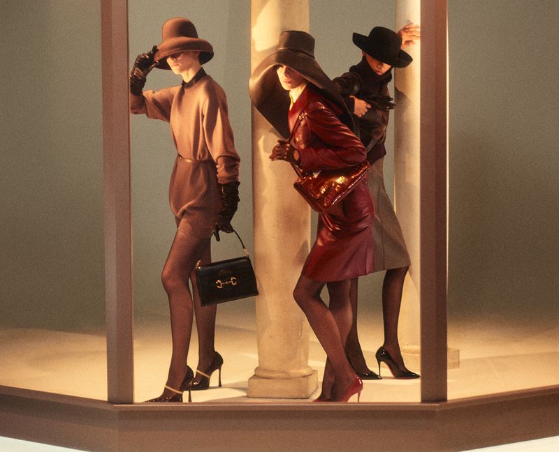 Three models wearing wide-brimmed hats and holding handbags pose between architectural columns in an editorial fashion shoot.