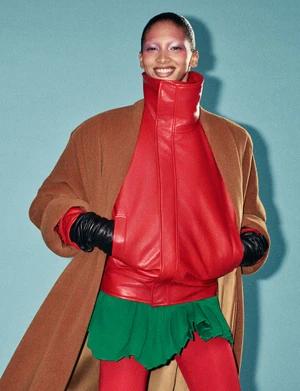 Model in brown coat, red jacket and green skirt posing against a blue background for AnOther Magazine.