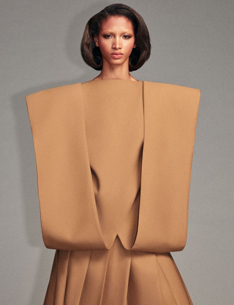 Model wearing a structured tan garment with architectural shoulders against a solid grey background