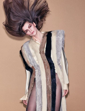 Model with long dark hair flying upward wearing a vertically striped fur vest against a textured tan background.