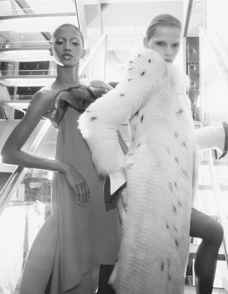 Two fashion models posing on a staircase, one wearing a fur coat and the other in a sleeveless dress.
