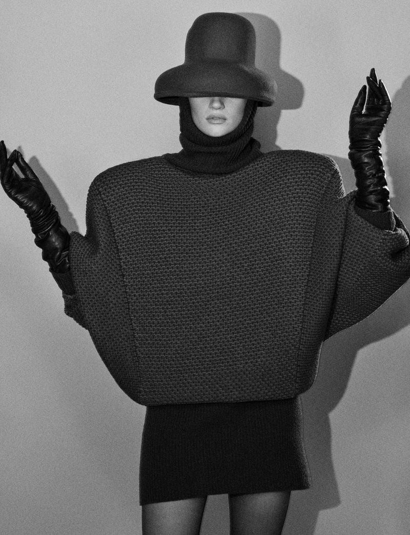 Monochrome fashion editorial image of a model wearing an oversized sculptural sweater, wide-brimmed hat, and leather gloves.