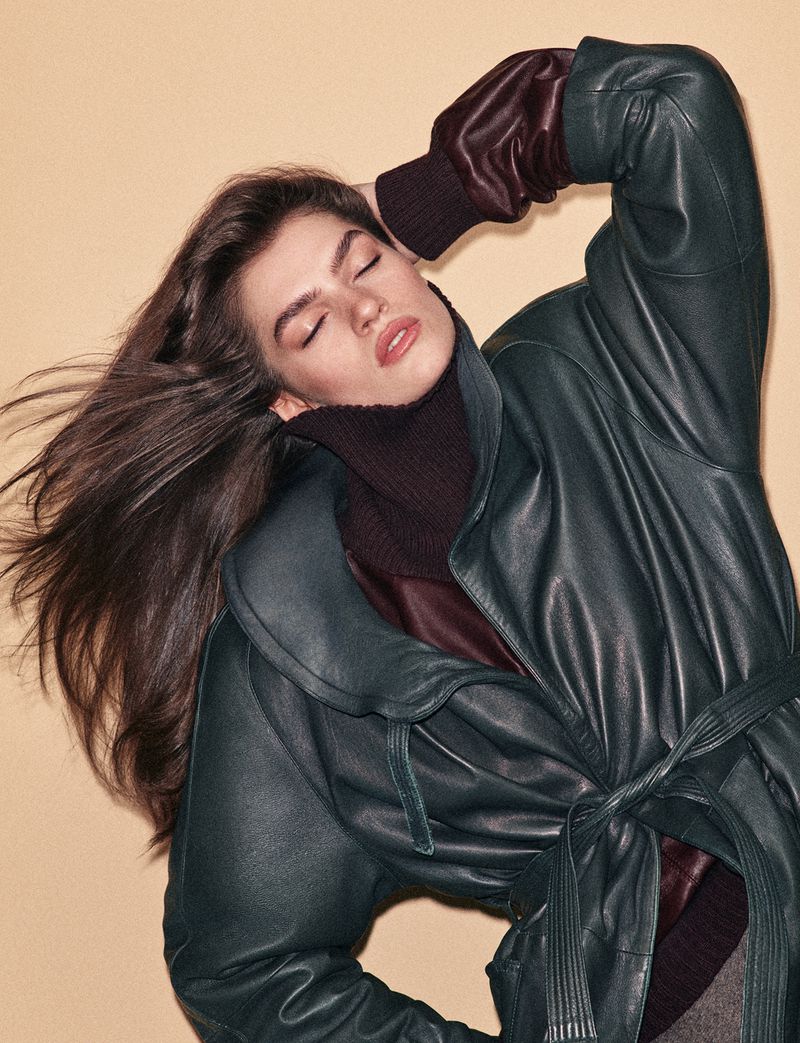Model with long dark hair wearing a green leather jacket and high-neck sweater against a plain neutral background.