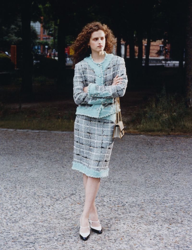 Model with curly red hair wearing a light blue and grey tweed skirt suit standing on a paved path outdoors.