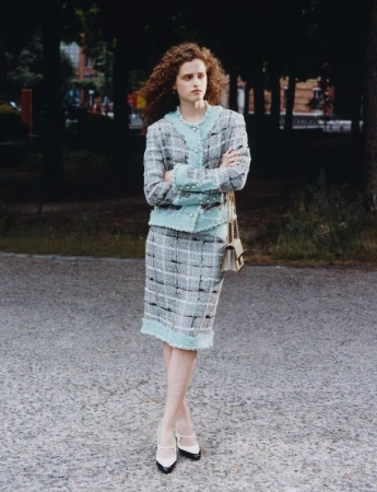 Model with curly red hair wearing a light blue and grey tweed skirt suit standing on a paved path outdoors.