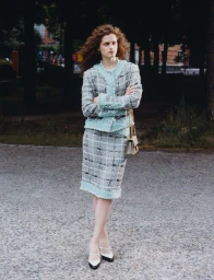 Model with curly red hair wearing a light blue and grey tweed skirt suit standing on a paved path outdoors.