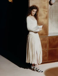 Model with long curly hair wearing a cream dress holding a book in a kitchen interior with wooden cabinets