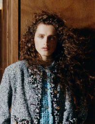 Model with long curly brown hair wearing a textured tweed jacket standing indoors against a wood panel wall