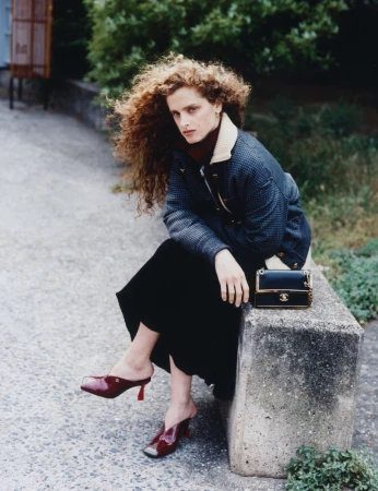 Model with voluminous curly hair sitting on a concrete ledge wearing a quilted jacket, dark skirt, and red mules