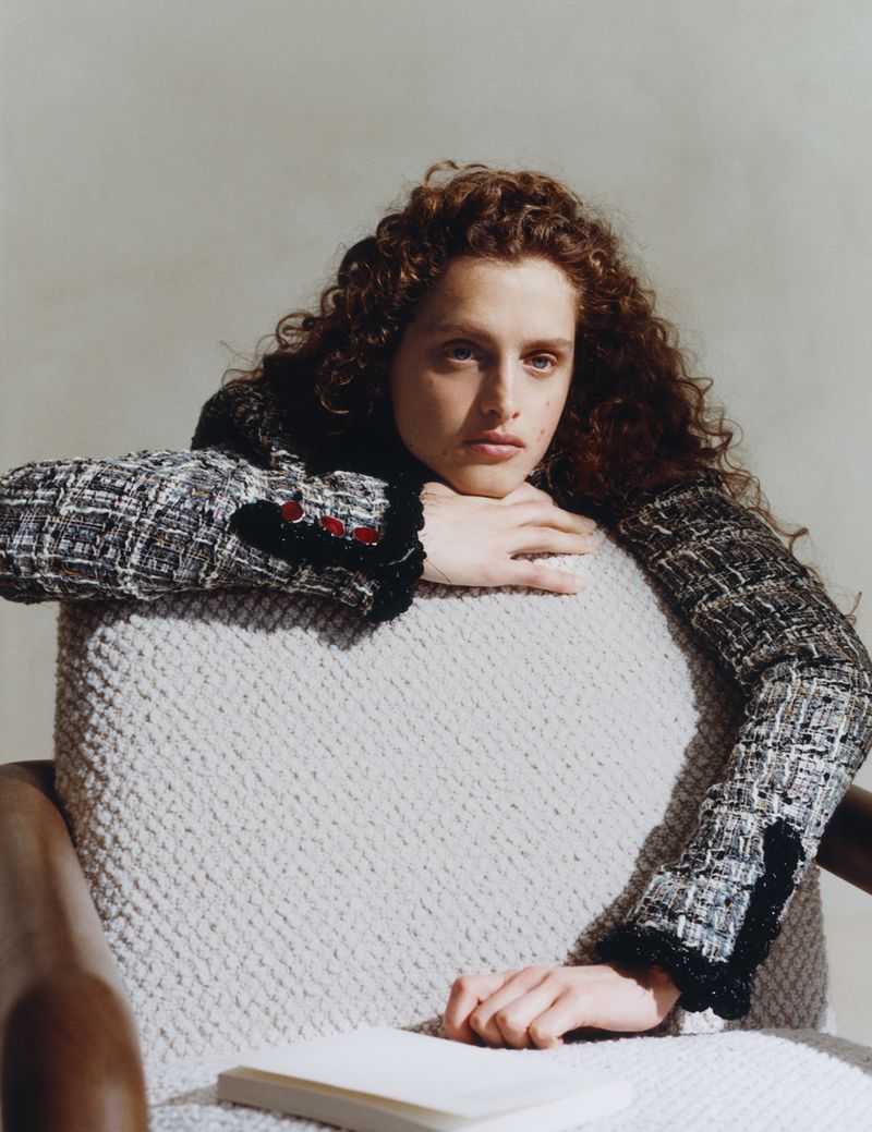 Model with curly brown hair wearing a textured tweed jacket seated and leaning over a textured armchair.