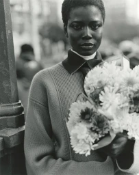 Black model holding a large bouquet of flowers wearing a collared sweater on a city street in a black and white fashion portrait.