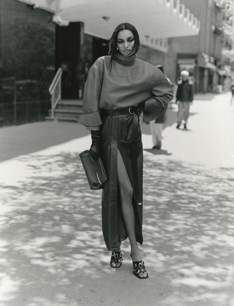Model standing on a city sidewalk wearing a high-neck top, a long skirt with a front slit, and holding a clutch bag.