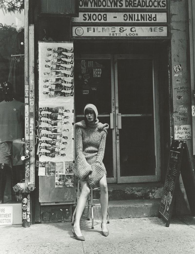 Model wearing a chunky turtleneck sweater and beanie, seated on a stool outside an urban storefront for 10 Magazine