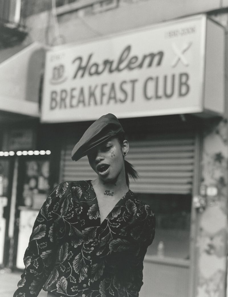 Monochrome fashion portrait of a model wearing a patterned top and beret under a Harlem Breakfast Club sign