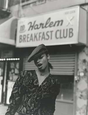 Monochrome fashion portrait of a model wearing a patterned top and beret under a Harlem Breakfast Club sign