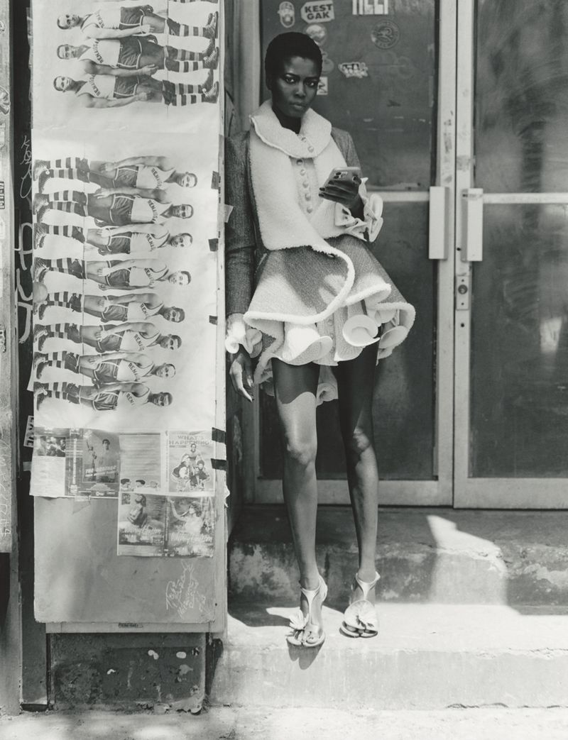 Model standing outdoors next to posters on a wall, wearing a textured jacket with white trim and strappy sandals.