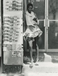 Model standing outdoors next to posters on a wall, wearing a textured jacket with white trim and strappy sandals.