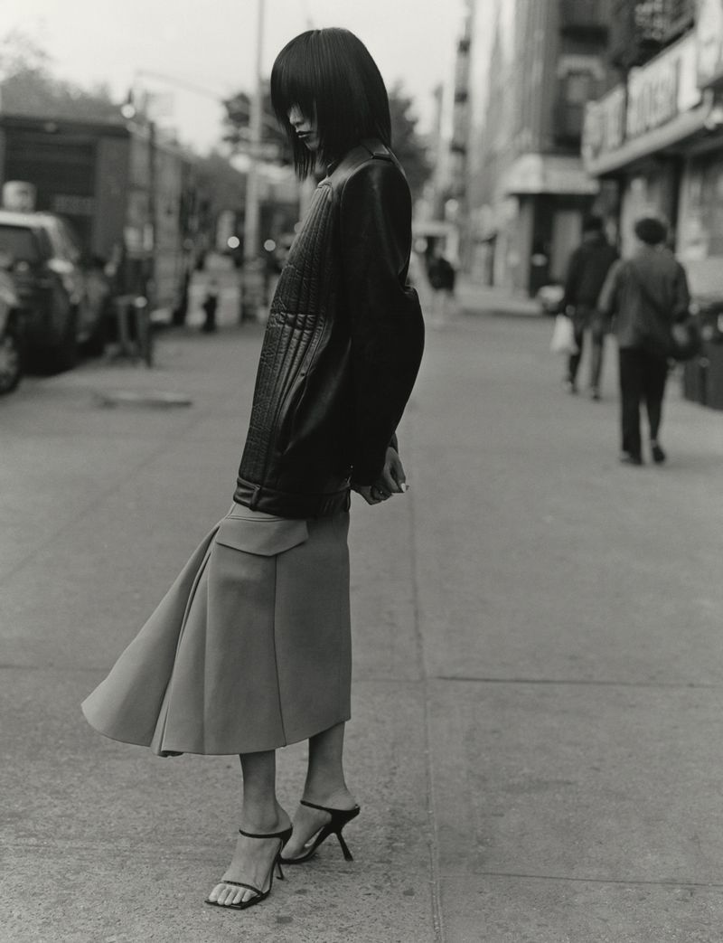 Model standing on a city street wearing a dark leather jacket and flared skirt, photographed in black and white.