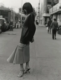 Model standing on a city street wearing a dark leather jacket and flared skirt, photographed in black and white.