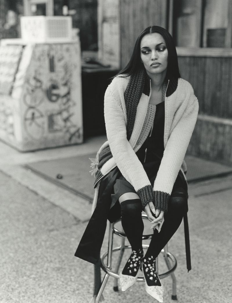 Model seated on a metal stool outdoors wearing a knit cardigan and patterned shoes in a black and white editorial photograph.
