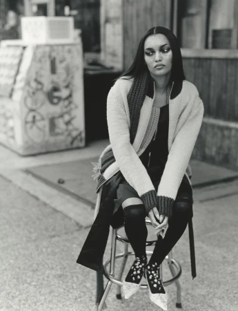 Model seated on a metal stool outdoors wearing a knit cardigan and patterned shoes in a black and white editorial photograph.