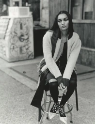 Model seated on a metal stool outdoors wearing a knit cardigan and patterned shoes in a black and white editorial photograph.