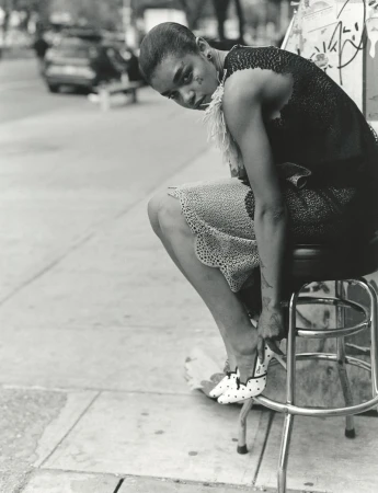 Black model wearing dark lace dress seated on a metal stool adjusting polka dot shoes on an urban street.