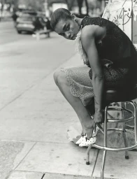Black model wearing dark lace dress seated on a metal stool adjusting polka dot shoes on an urban street.
