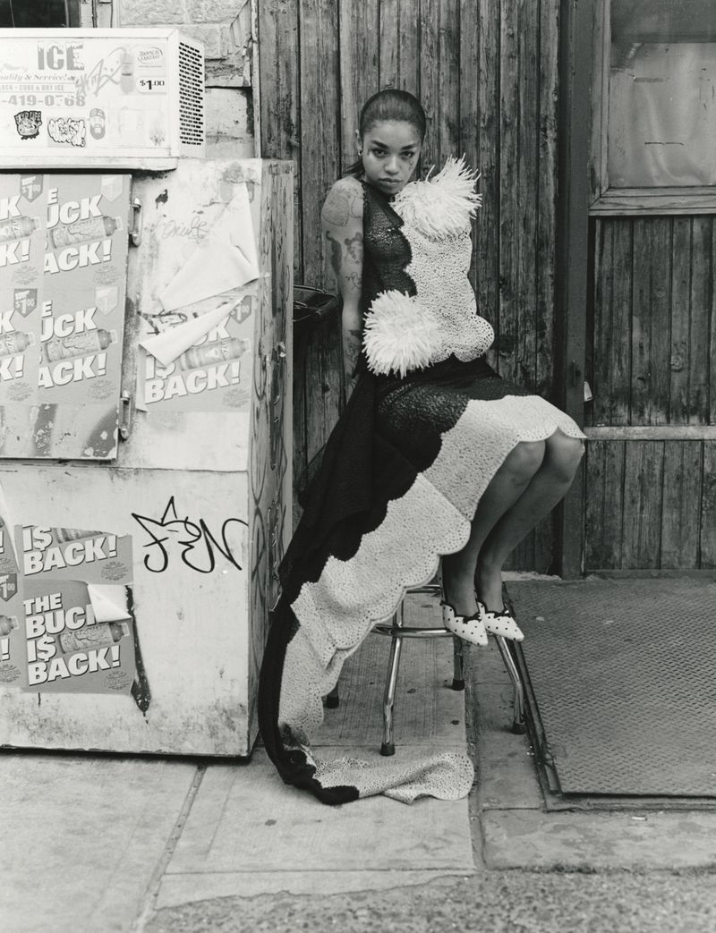Model seated on a stool next to a graffitied vending machine wearing an ornate textured dress.