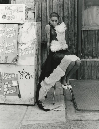 Model seated on a stool next to a graffitied vending machine wearing an ornate textured dress.