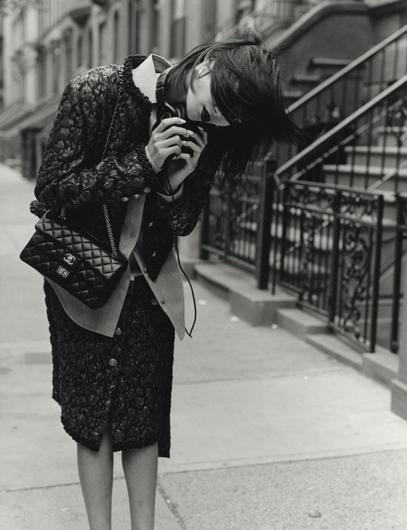 Model in black textured jacket and skirt holding a camera on a city sidewalk with stoop stairs