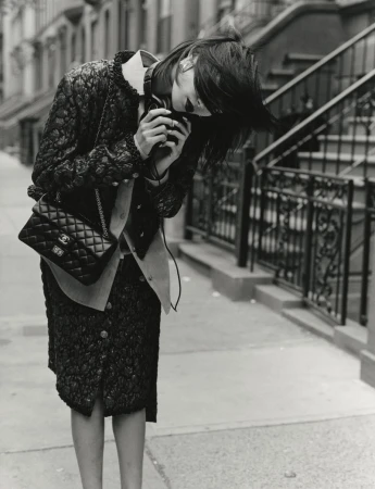 Model in black textured jacket and skirt holding a camera on a city sidewalk with stoop stairs