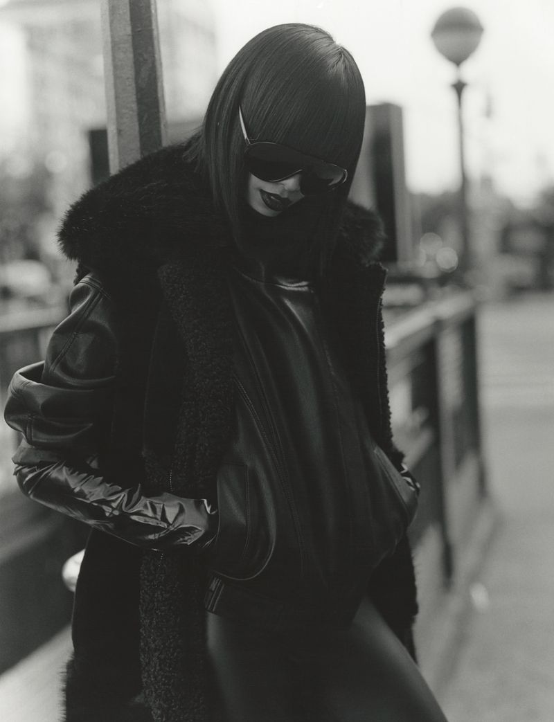Model wearing dark sunglasses and heavy black outerwear posing outdoors in a monochrome urban scene