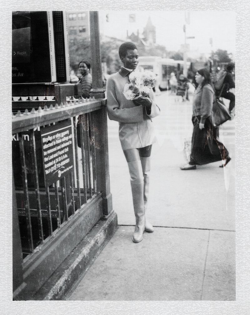 Black model standing on a city sidewalk holding flowers near a subway entrance in a black and white fashion editorial photograph.