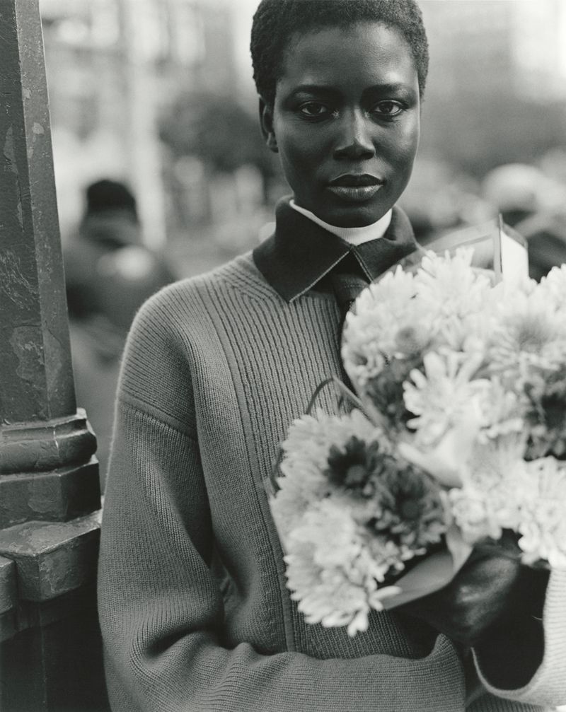 Black model holding a large bouquet of flowers wearing a collared sweater on a city street in a black and white fashion portrait.