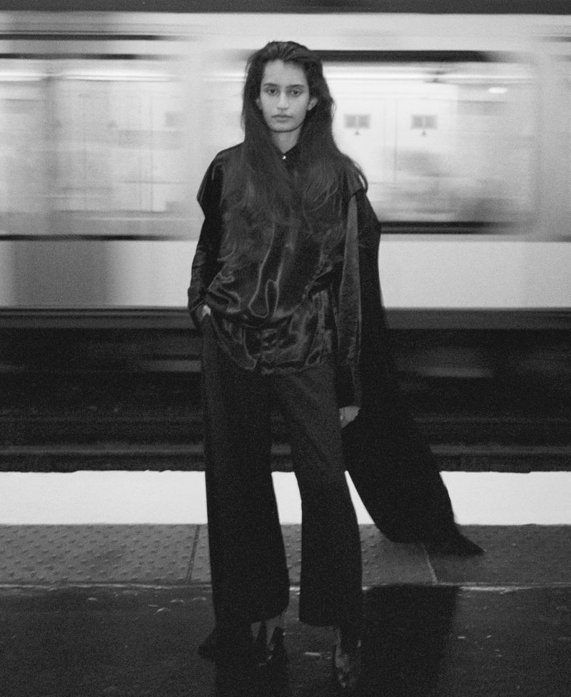 Monochrome fashion editorial image of a model standing on a subway platform with a blurred train passing behind.