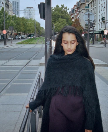 Woman in a black fringed cape standing beside tram tracks on a city street