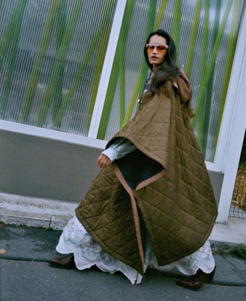 Model in quilted brown cape and patterned skirt posing outdoors against a translucent ribbed structure
