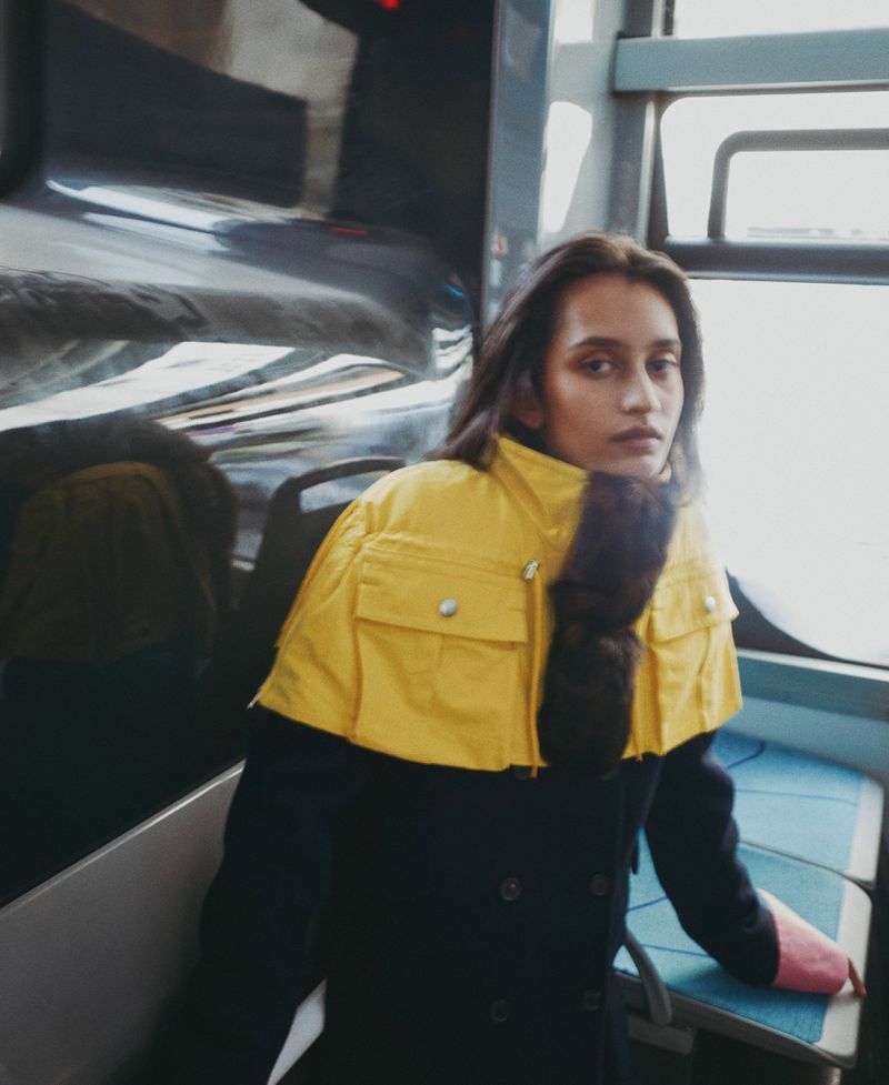Model wearing a colorblocked yellow and dark coat seated inside a train near a large window