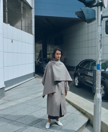 Model wearing a grey hooded cape coat standing on a concrete city street with parked cars and a traffic light pole.