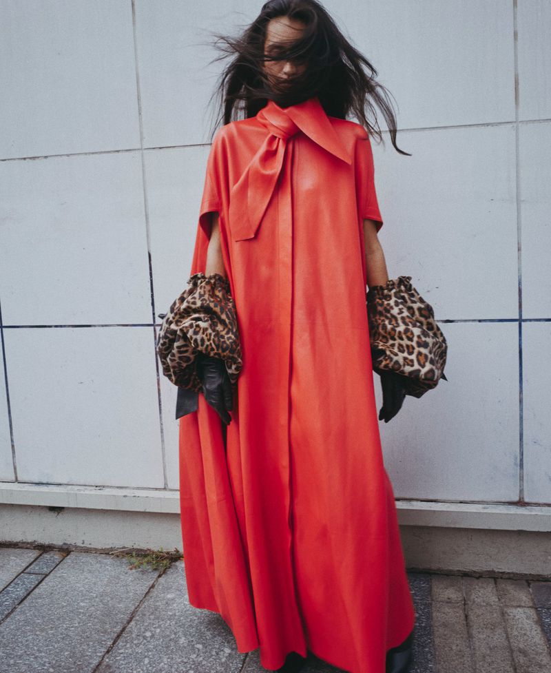Model wearing a vibrant red cape gown and leopard print sleeves standing against a tiled exterior wall.
