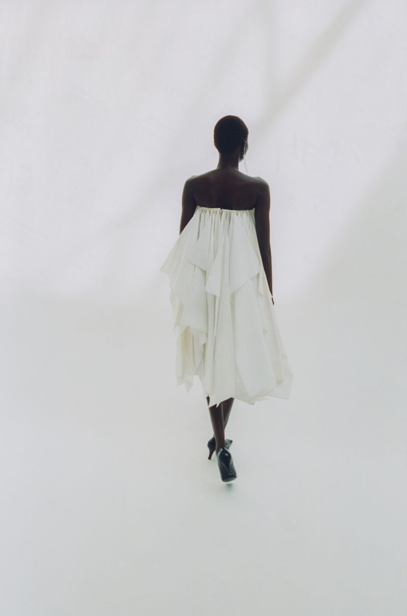 A Black woman seen from behind wearing a white strapless pleated dress and dark heels against a bright white background.