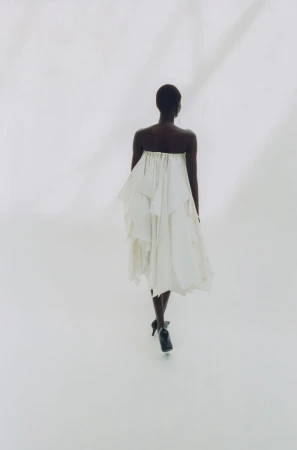 A Black woman seen from behind wearing a white strapless pleated dress and dark heels against a bright white background.