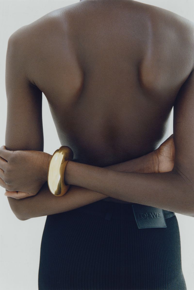 Close-up back view of a person wearing a chunky gold bracelet and dark skirt with arms crossed against a plain light background