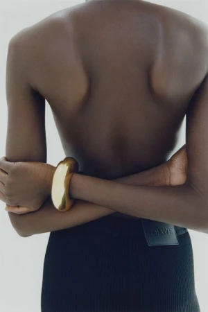 Close-up back view of a person wearing a chunky gold bracelet and dark skirt with arms crossed against a plain light background