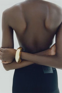 Close-up back view of a person wearing a chunky gold bracelet and dark skirt with arms crossed against a plain light background
