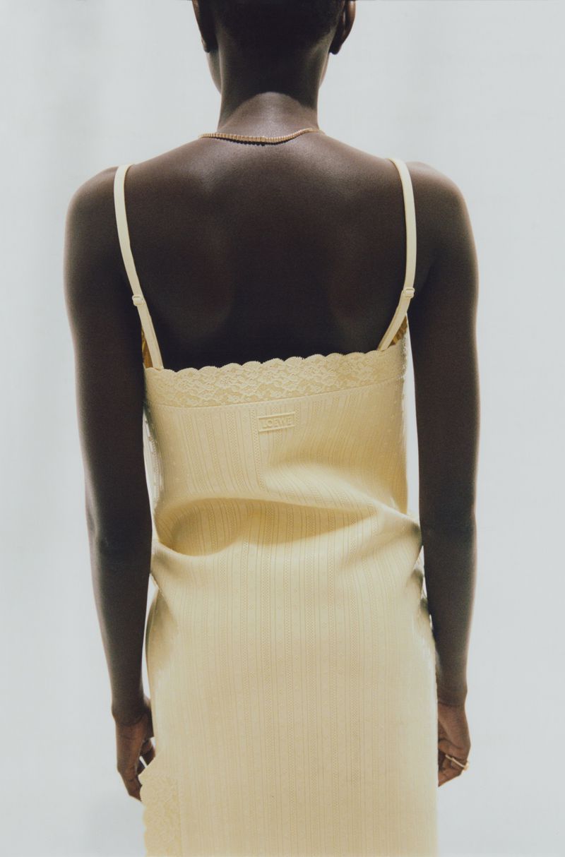 Rear view of a Black woman wearing a yellow slip dress and gold necklace against a seamless grey background.